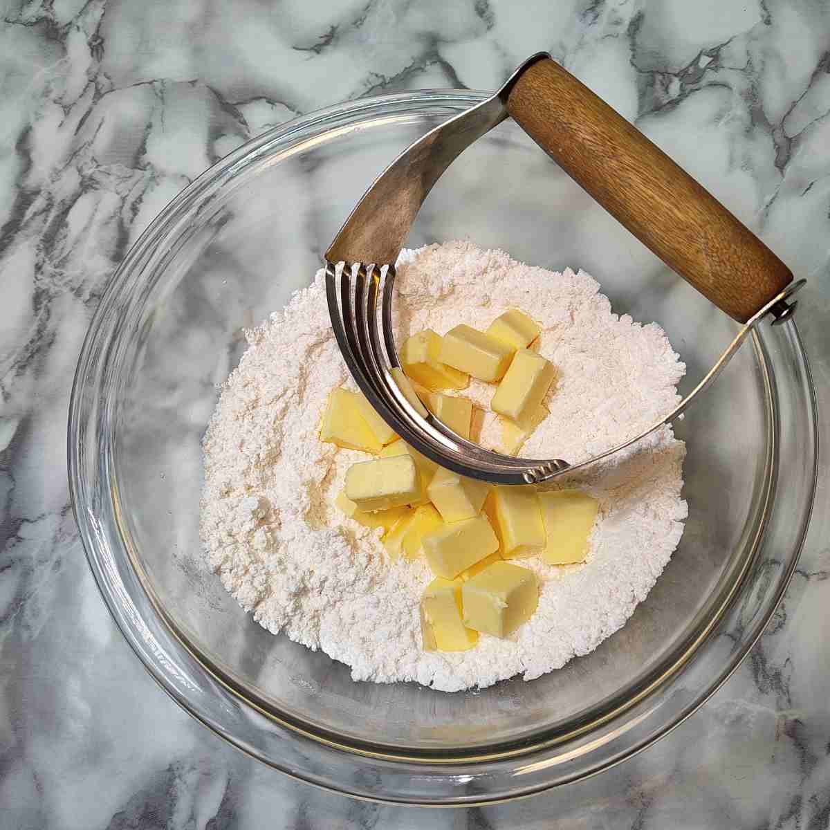 Cubes of yellow butter and the dry ingredients in a clear glass bowl with a blue whisk.