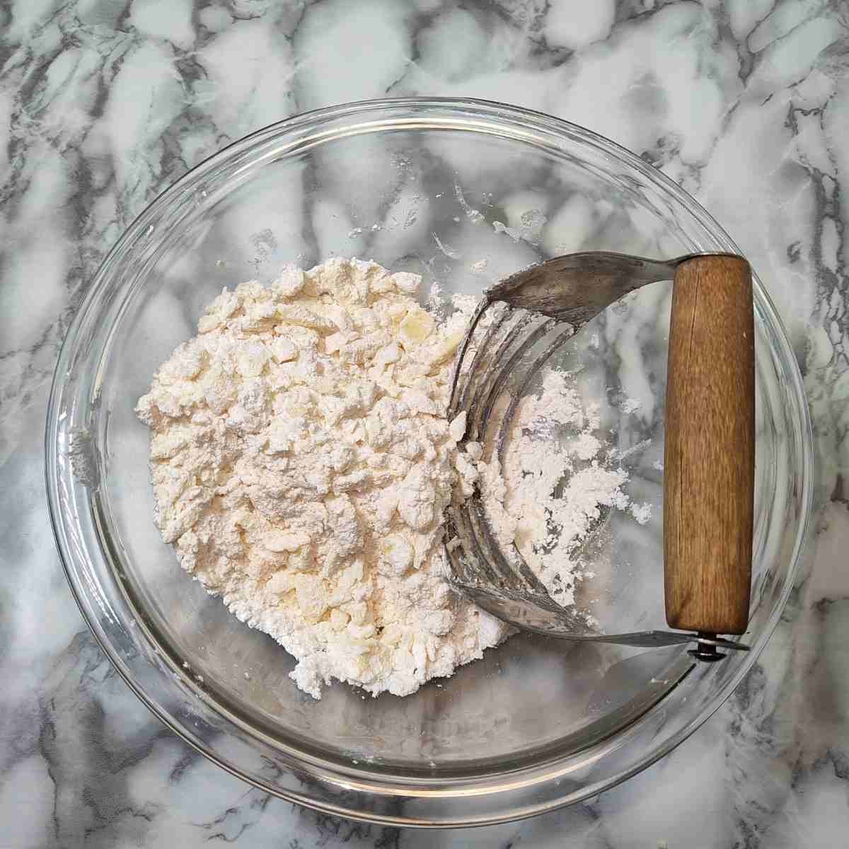 A clear glass bowl with lumpy, white flour and a brown handled pastry cutter.