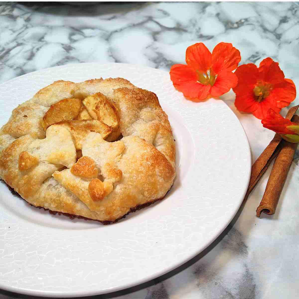 A light brown galette on a white plate with orange flowers and cinnamon sticks.