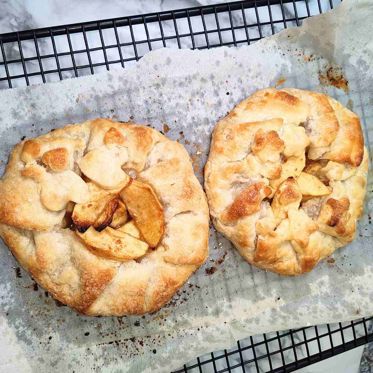 Two biege, yellow, and brown galettes cooling on oily parchment paper on a cooling rack.