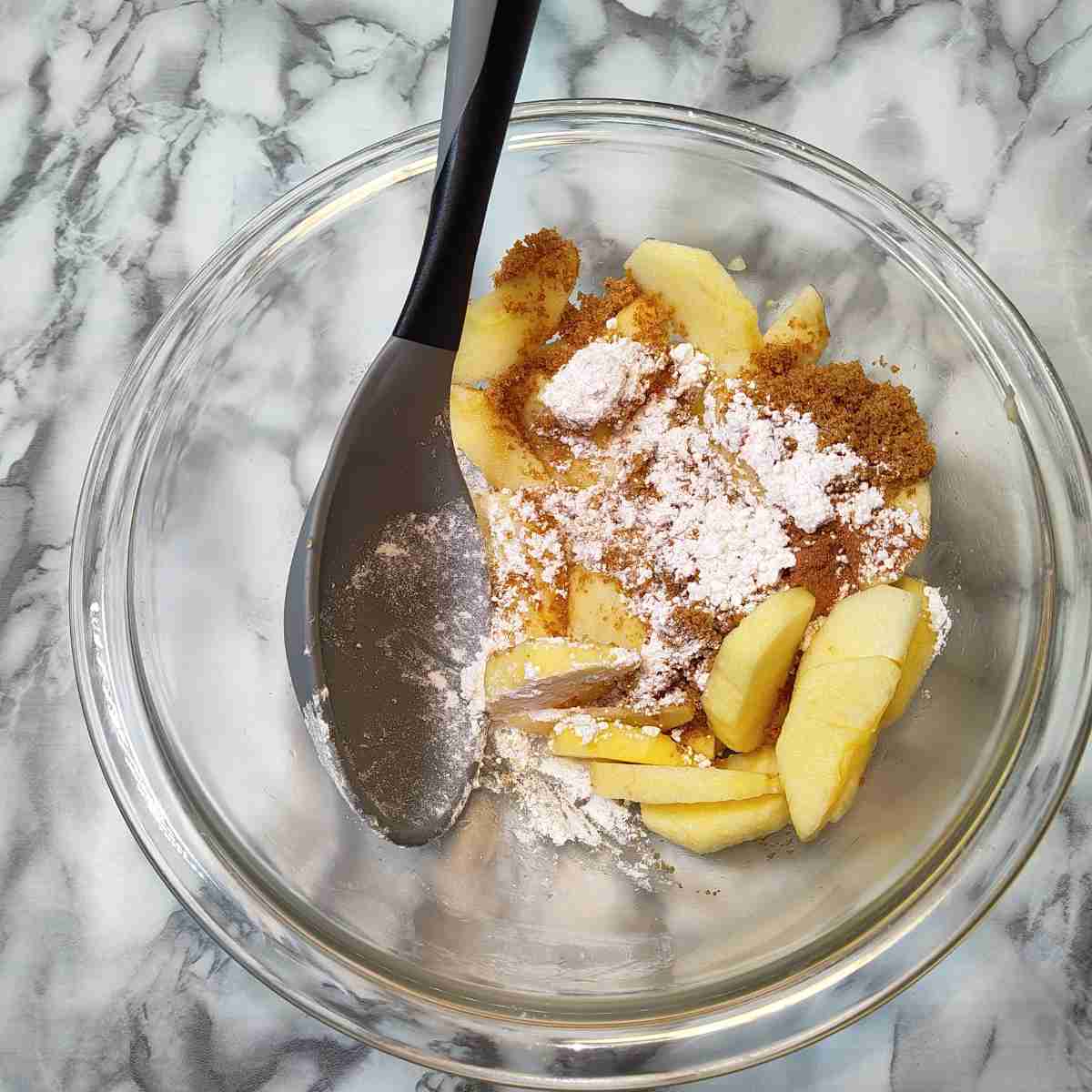 Light yellow apple slices with brown sugar and white flour ready for mixing in a clear bowl with a grey spoon.
