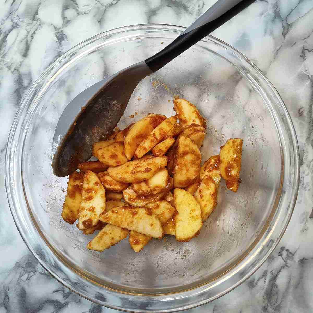 Sliced apples covered in brown sugar and cinnamon in a clear bowl with a grey spoon.