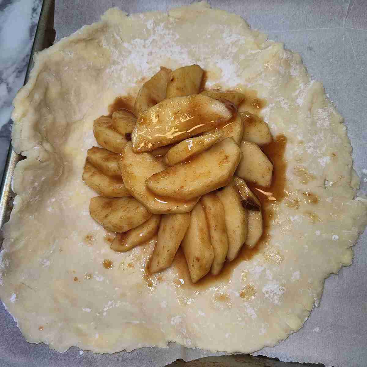 One beige galette rolled flat with brown juicy apple slices arranged in the middle on a lined cookie sheet.