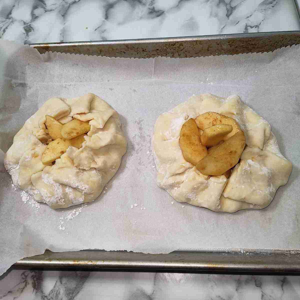 Two uncooked white galettes folded over the yellow and brown apple filling on a parchment paper lined cookie sheet.