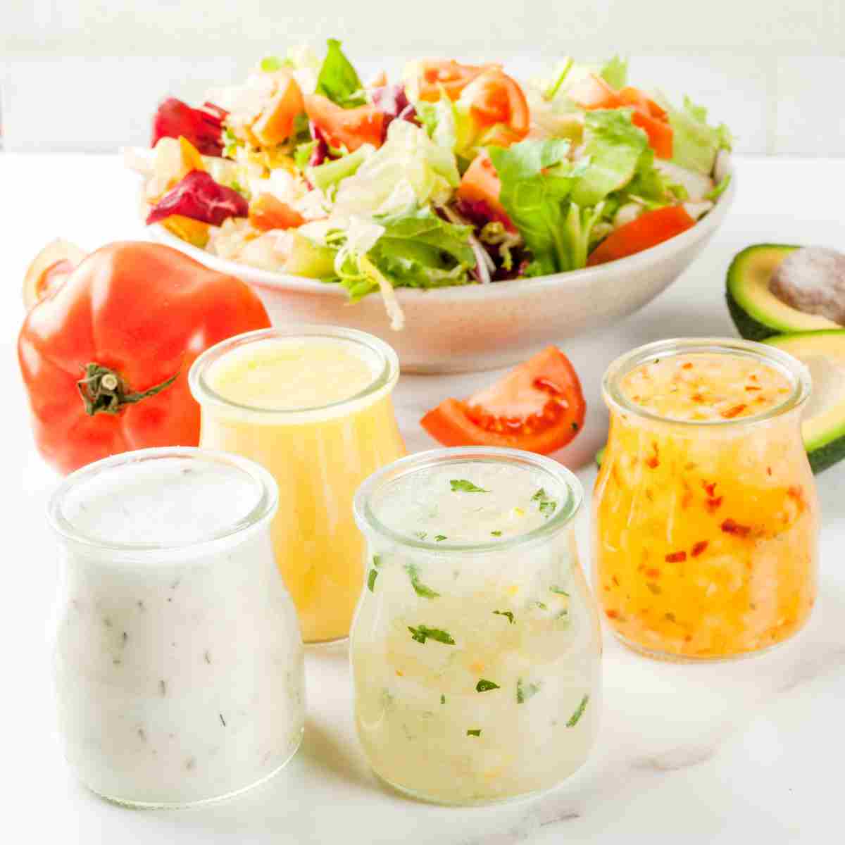 A bowl of colorful salad, whole red tomato, and one wedge, one green and yellow avocado sliced in half, and four different collors of jars of salad dressing.