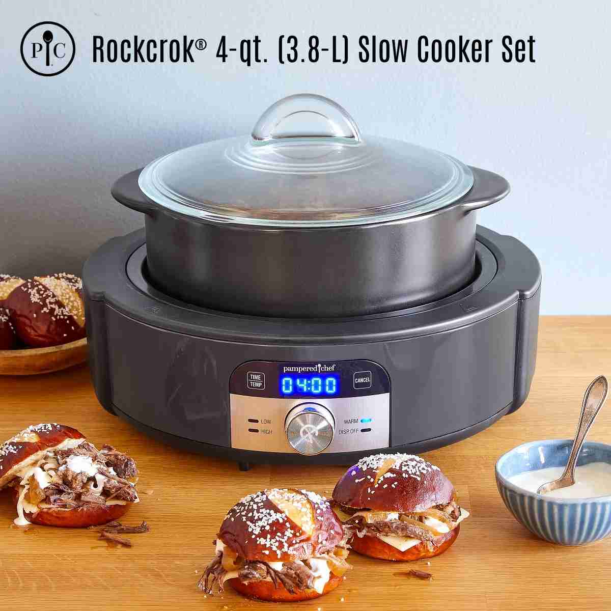 A black two piece slow cooker with a clear lid surrounded by brown meat in reddish-brown buns and bowl of white sauce.