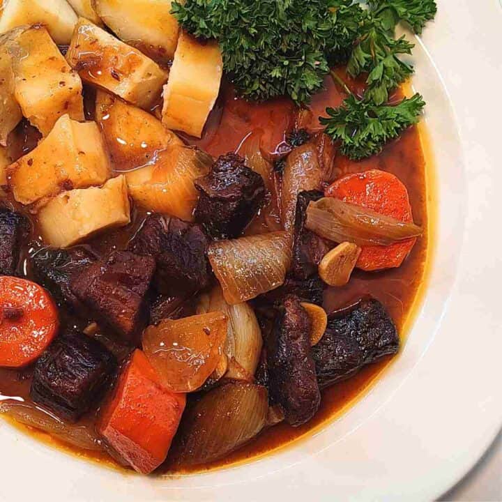 A white dinner plate with orange, beige, and dark brown meat with brown gravy, yellow potato chunks, and green parsley.