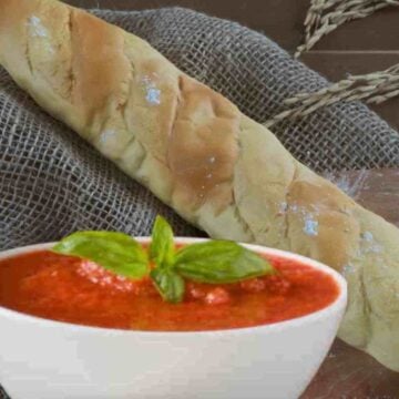 White bowl of red tomato sauce with a green basil sprig with beige and brown baguette.