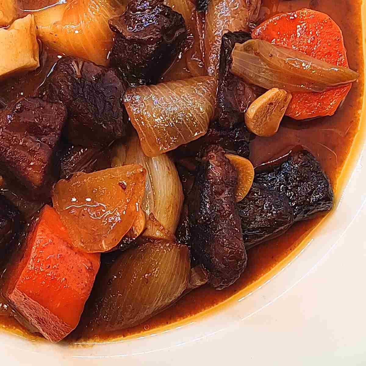 White plate with cutup pieces of orange carrots, dark brown meat, beige onions and garlic with brown gravy.