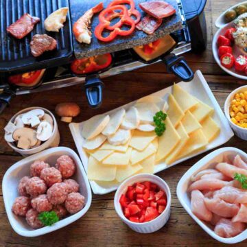 A black grill called a raclette with various kinds of food cooking on it and around it in white dishes.