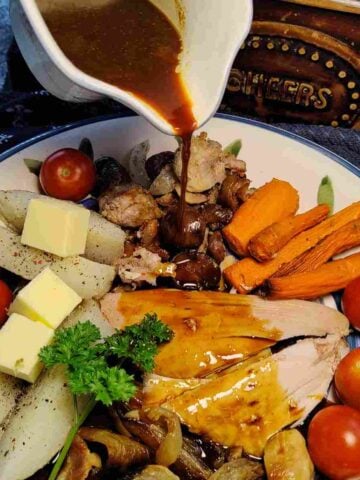 Pouring dark gravy on a plate of sliced turkey dinner and colorful vegetables.