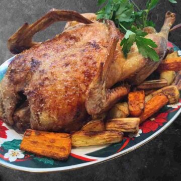 A cooked golden brown duck surrounded by cooked orange and yellow vegetables and green parsley on a colorful oval plate.