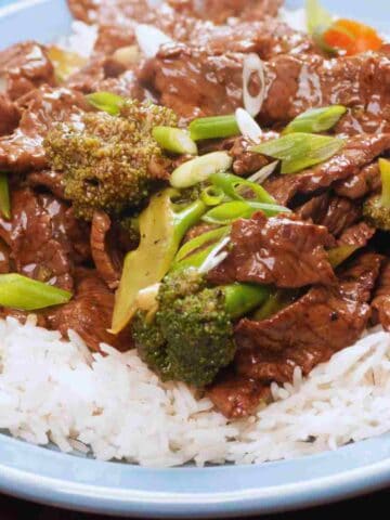 A light blue plate filled with sliced brown beef steak in a sauce with green vegetables on a bed of white rice.