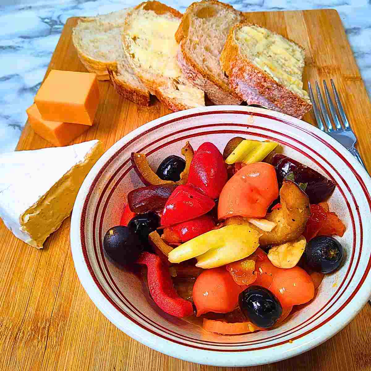 A bowl of colorful vegetables on a wooden board with sliced bread and a variety of cheeses.