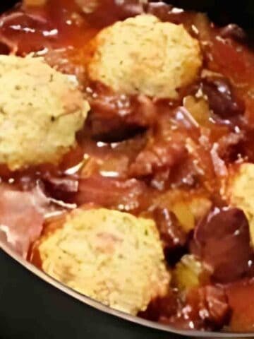 A black pot of reddy-brown venison meat chunks covered in reddish gravy with white dumplings spotted with green thyme.