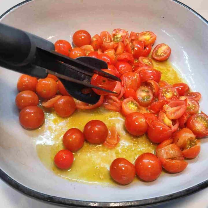 Red cherry tomatoes cooking in oil in a pot and being sliced with kitchen scissors.