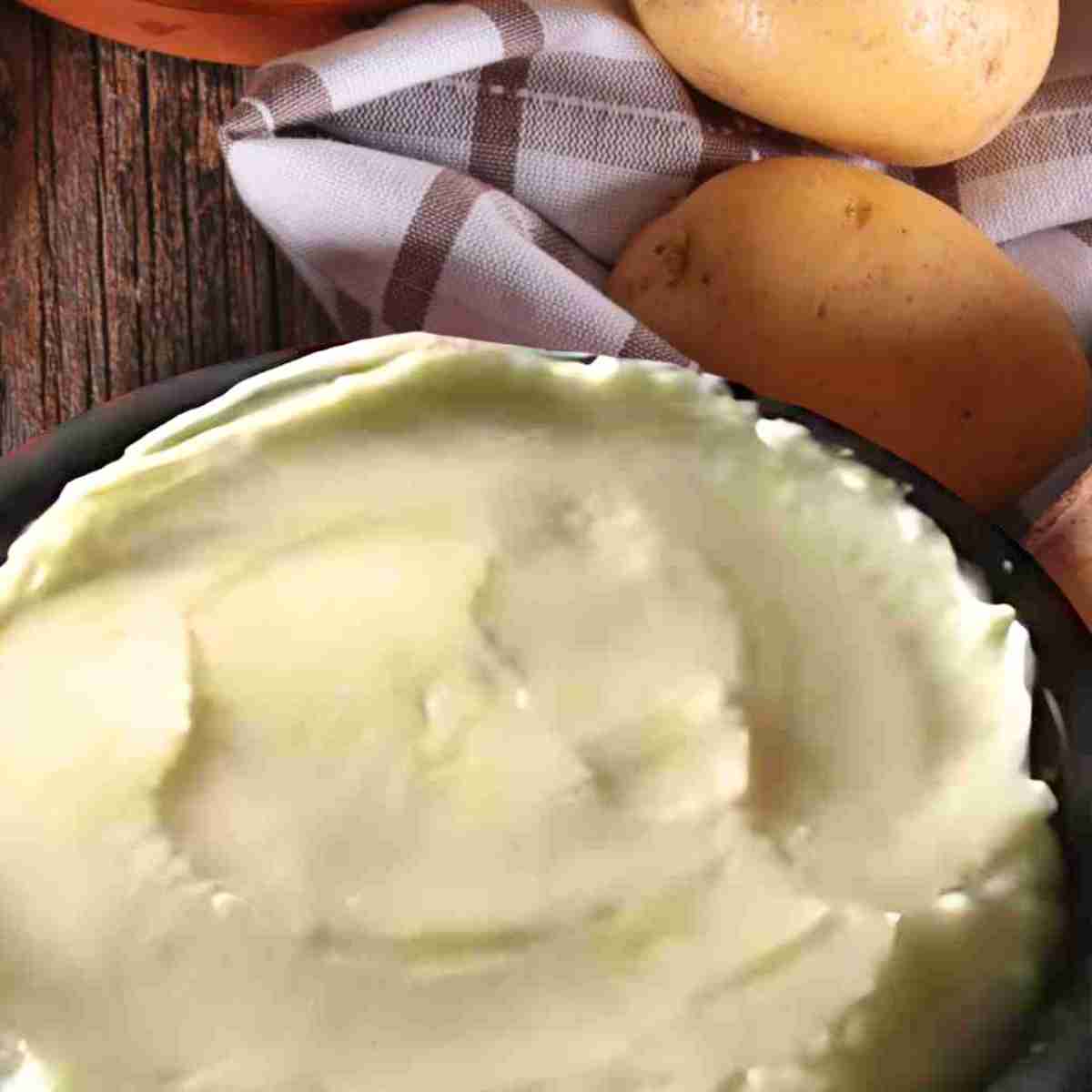 A bowl of white mashed potatoes surrounded by beige potatoes on a brown plaid tea towel.