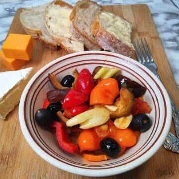 A bowl of colorful vegetables on a wooden board with sliced bread and a variety of cheeses.