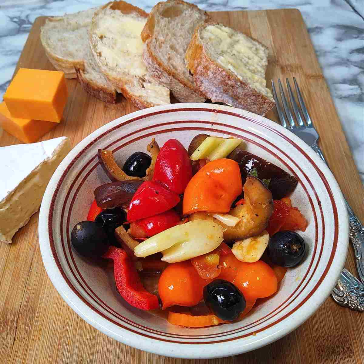 A bowl of colorful vegetables on a wooden board with sliced bread and a variety of cheeses.