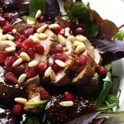 Pork loin roast sliced and topped with figs, pomegranate seeds, and pine nuts on a plate of greens.