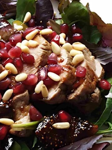 Pork loin roast sliced and topped with figs, pomegranate seeds, and pine nuts on a plate of greens.